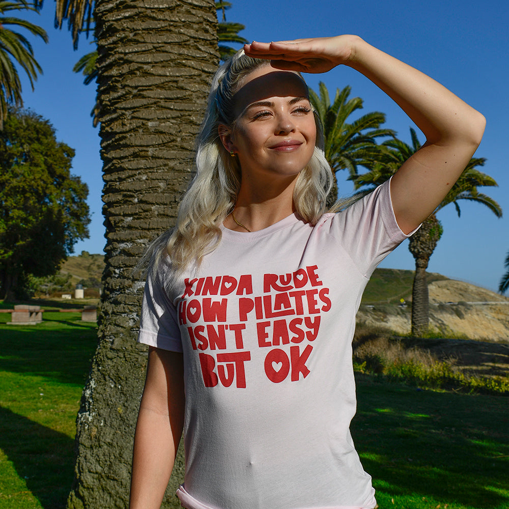 Woman wearing a pink unisex t-shirt that says "Kinda Rude how pilates isn't easy but ok"