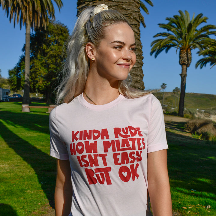 Woman wearing a pink unisex t-shirt from The Movement Shop that says "Kinda Rude how pilates isn't easy but ok"
