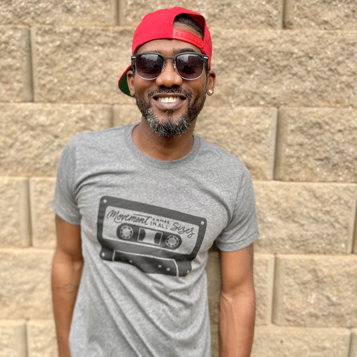 Man wearing a deep heather unisex t-shirt that says "Movement Comes In All Sizes" designed within a mixed tape