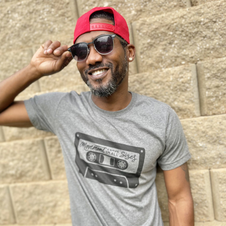 Man wearing a deep heather unisex t-shirt that says "Movement Comes In All Sizes" designed within a mixed tape