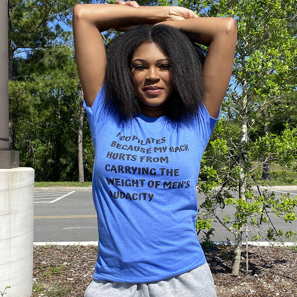 Woman wearing a blue t-shirt that says "I do Pilates because my back hurts from carrying the weight of men's audacity". T-Shirt is from The Movement Shop