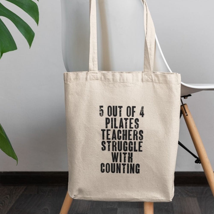 Canvas tote bag that is on the back of a white chair. Tote bag says "5 out of 4 Pilates Teachers Struggle With Counting" on one side.