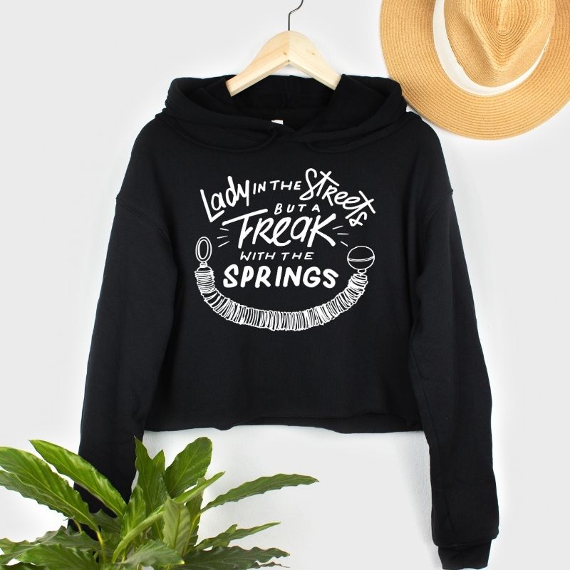 Black Women's Cut Crop Fleece Hoodie That says Lady in The Streets But A Freak With The Springs in White text. There is a design of a pilates spring on the bottom.