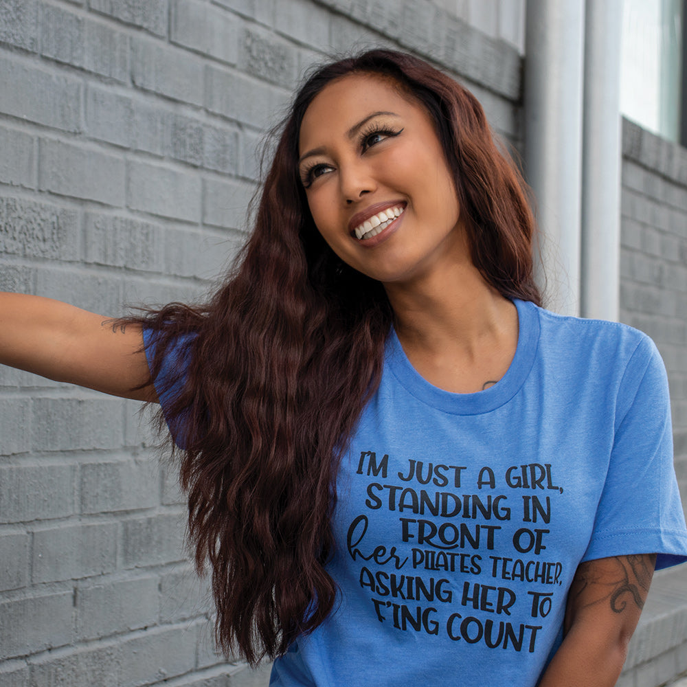 A woman wearing a heather columbia unisex crew t-shirt that says "I'm just a girl, standing in front of her pilates teacher , asking her to f'ing count"