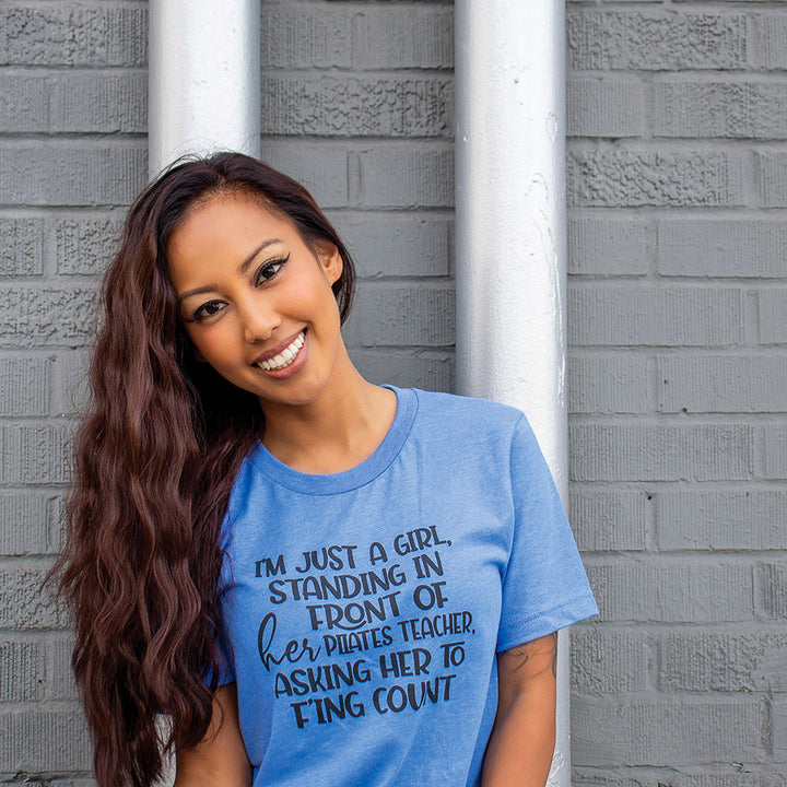 A woman wearing a heather columbia unisex crew t-shirt that says "I'm just a girl, standing in front of her pilates teacher , asking her to f'ing count"