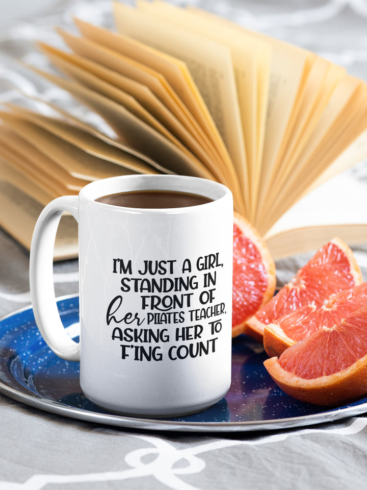 15 oz white coffee mug that says "I'm just a girl, standing in front of her Pilates Teacher, asking her to f'ing count". There is a book in the background.