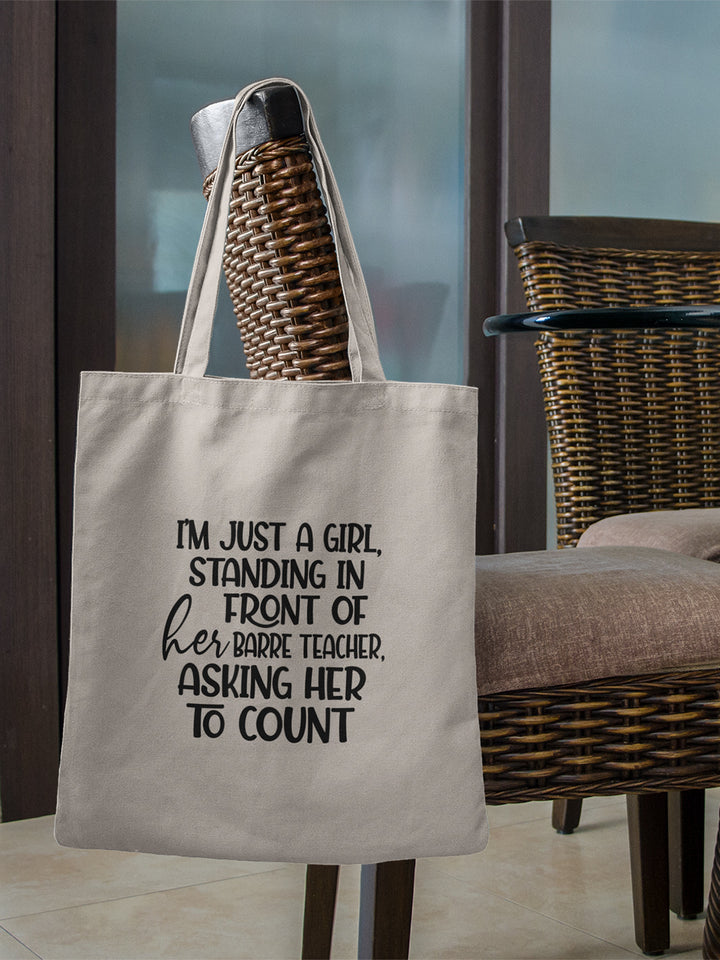 100% cotton canvas tote bag with handles that says "I'm just a girl, standing in front of her Barre Teacher, asking her to count". Bag is hanging off the back a wicker dining room chair.