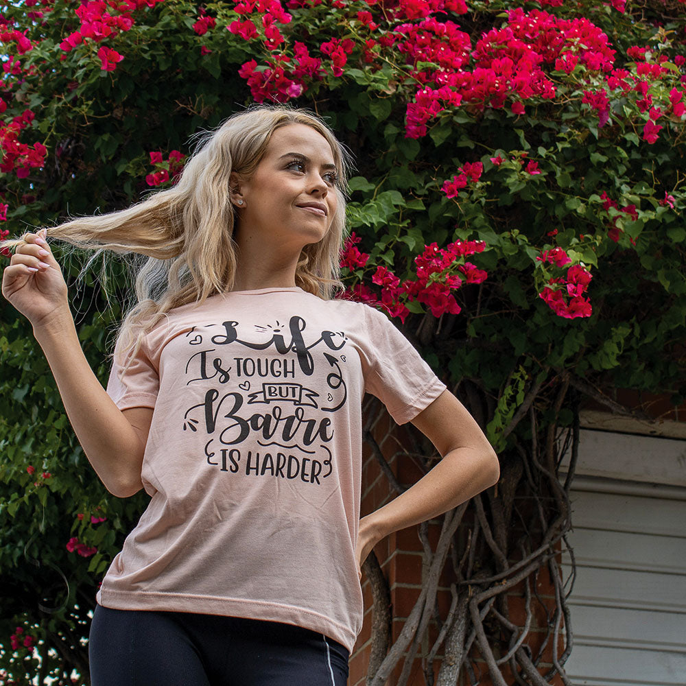 Woman wearing a heather peach unisex crew-t-shirt that says "Life is Tough But Barre Is Harder".