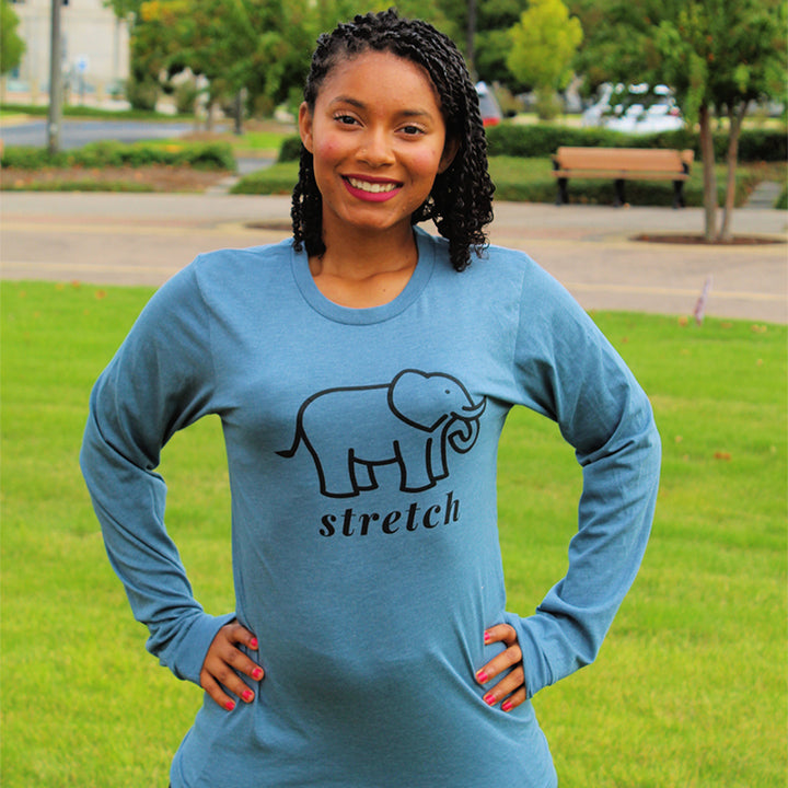 Woman wearing a long sleeve pilates shirt that is heather deep teal color and has an elephant in black outline and says the word "stretch" in black text