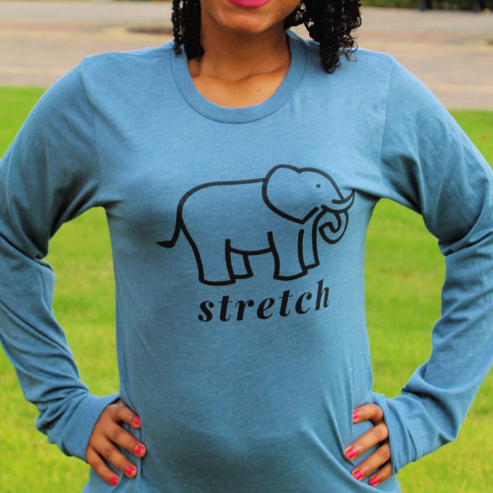 Woman wearing a long sleeve pilates shirt that is heather deep teal color and has an elephant in black outline and says the word "stretch" in black text