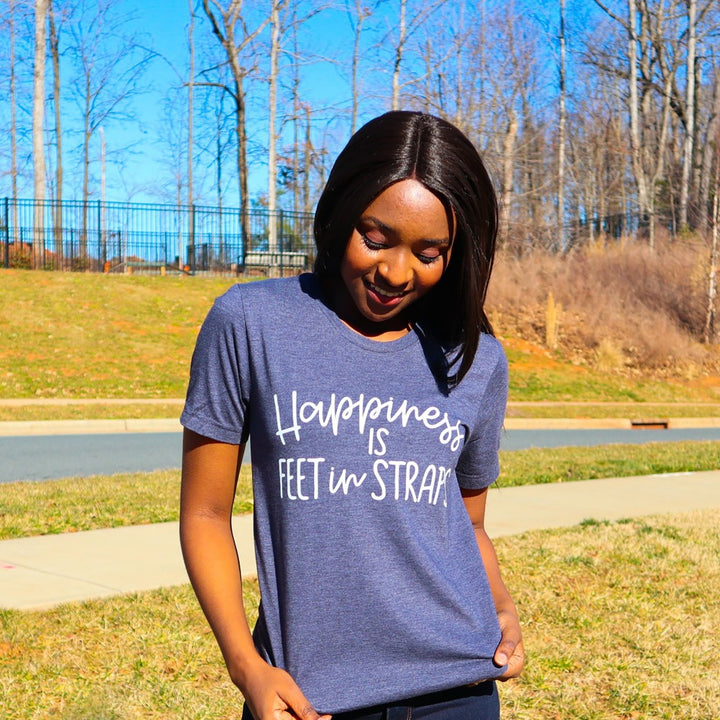 A woman wearing a heather navy unisex crewneck t-shirt that says "happiness is feet in straps" in white script text