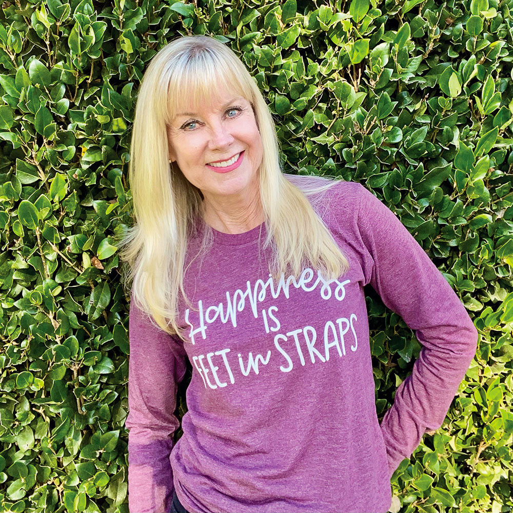 A woman wearing a long sleeve unisex crewneck long sleeve maroon triblend shirt that reads "happiness is feet in straps" in white script text
