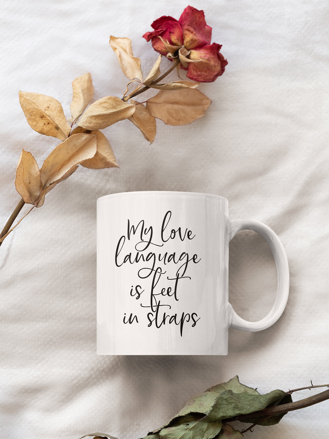 White 11oz coffee mug against a white background with dried roses that said "My Love Language Is Feet In Straps" black script