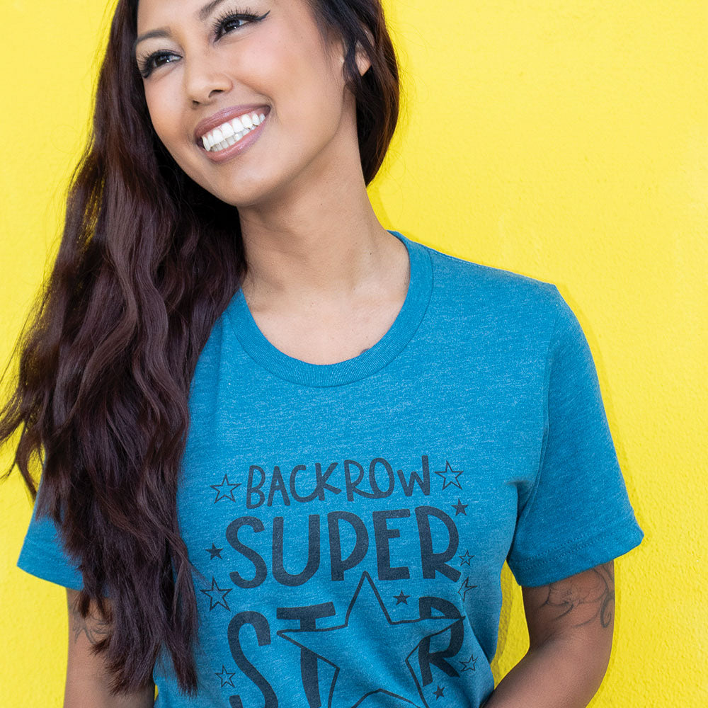 Woman in front of a bright yellow background wearing a Teal unisex crewneck t-shirt that says "Back Row Super Star"