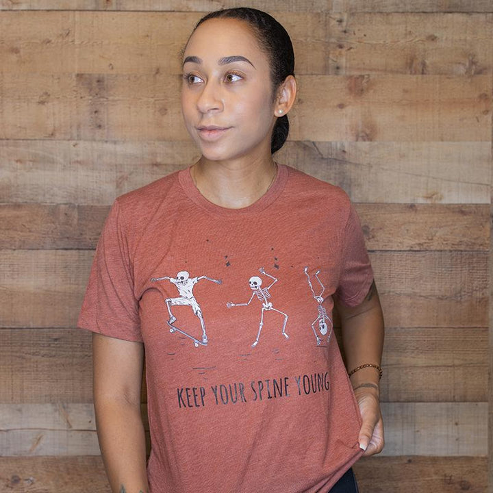 Woman wearing a heather orange unisex crewneck t-shirt that has three skelatons dancing. Text under the skelatons says Keep Your Spine Young in black text.