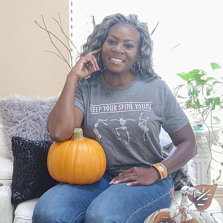 Woman wearing a deep heather grey unisex crewneck t-shirt that says "Keep Your Spine Young" in white with skeletons dancing