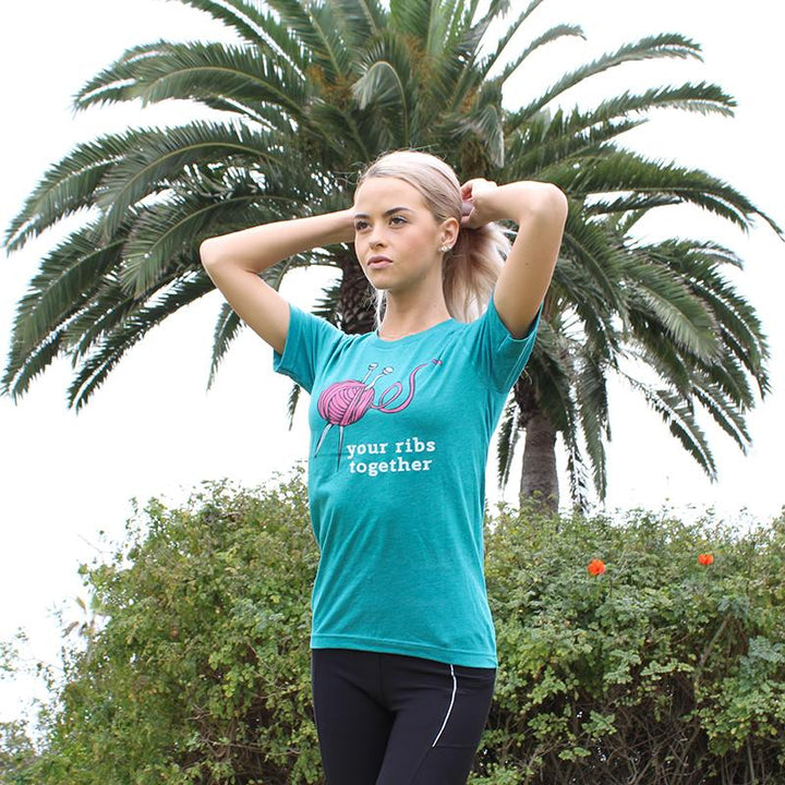 Woman wearing a deep heather teal unisex Crewneck T-Shirt that has a deep purple knitting needles and ball of yarn with the words "your ribs together:" in white text.