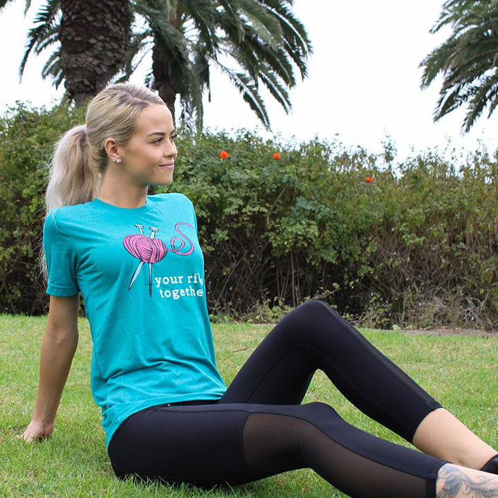 Woman wearing a deep heather teal unisex Crewneck T-Shirt that has a deep purple knitting needles and ball of yarn with the words "your ribs together:" in white text.