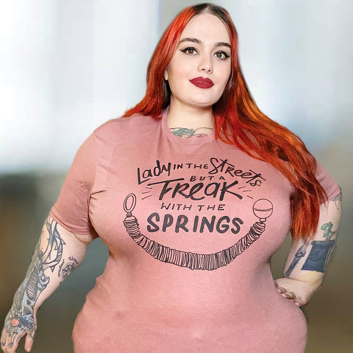 Woman wearing a mauve crew neck t-shirt that says "Lady in the streets but a freak with the springs" and a Pilates Reformer Spring design