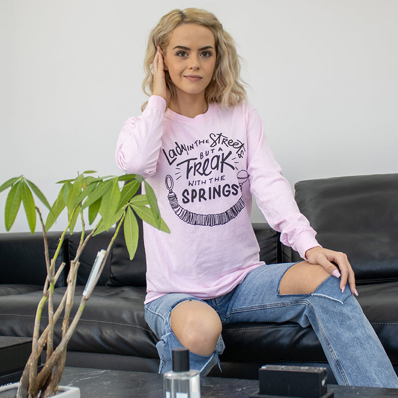 A Woman Wearing a unisex crew neck long sleeve shirt that is pink and says 'Lady In The Streets But A Freak With The Springs" in black text and black pilates spring under the text