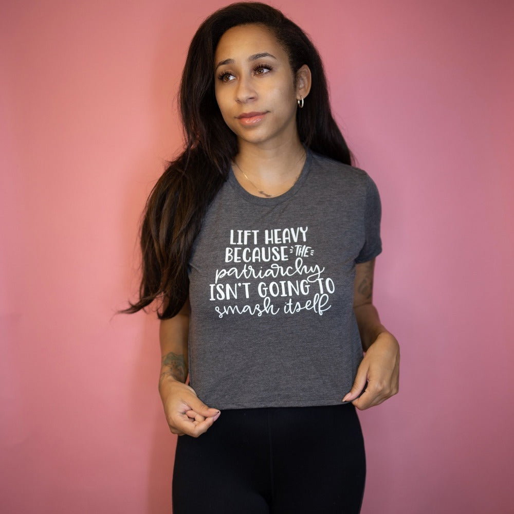 Woman wearing a deep heather crop top tshirt that says "Lift Heavy Because The Patriarchy Isn't Going To Smash Itself". Woman is leaning against a pink background.