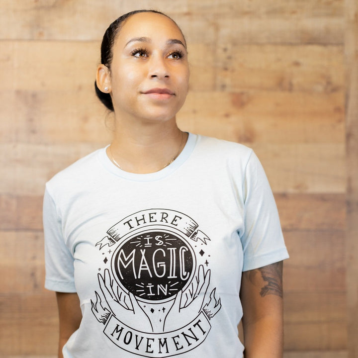 woman wearing heather ice blue crewneck t-shirt that says "There Is Magic In Movement" in black text. The words "is Magic In" is designed to look like a crystal ball.
