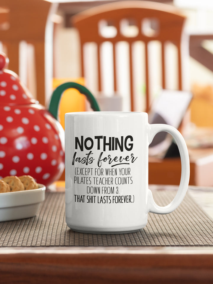 15oz Mug that says "Nothing Lasts Forever (Except For When You Pilates Teacher Counts Down From 3. That Shit Lasts Forever.)". The mug is sitting on a table with a chair in the background.