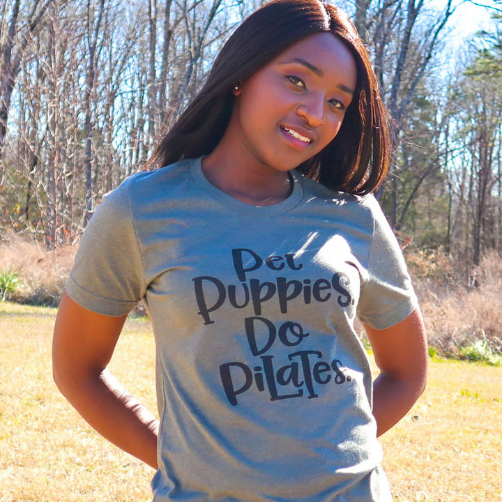 Woman wearing a unisex crewneck green t-shirt that says "Pet Puppies, Do Pilates" in black text