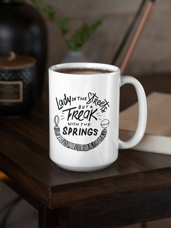 15oz Coffee Mug that says Lady In The Streets But A  Freak With The Springs. There is Pilates reformer spring under the text. Design is in black. Mug is sitting on a dark wooden table.