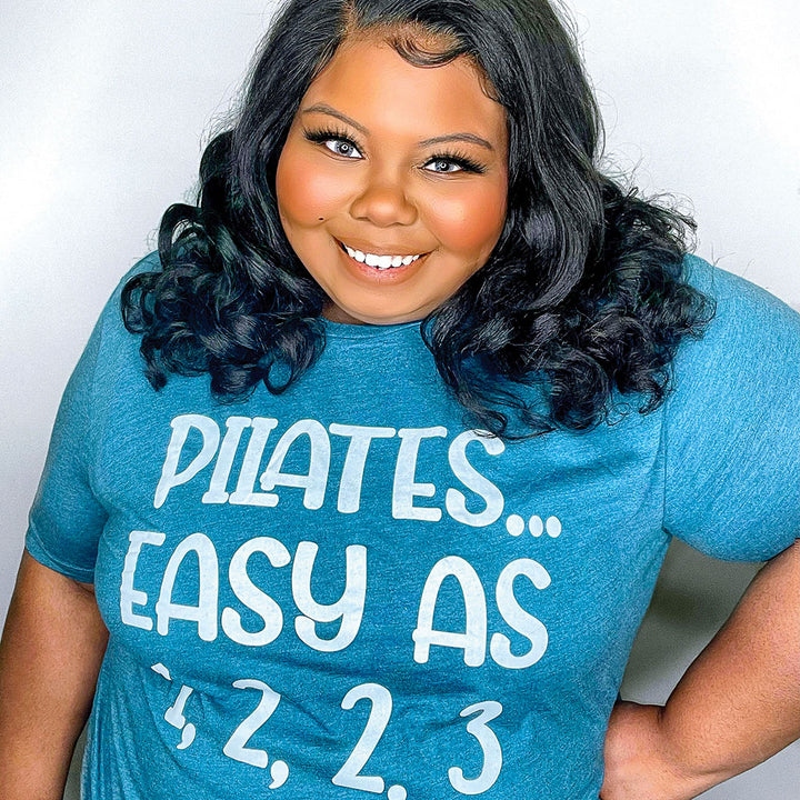 Woman wearing a heather deep teal unisex crewneck t-shirt that says "Pilates easy as 1,2,2,3" in white text