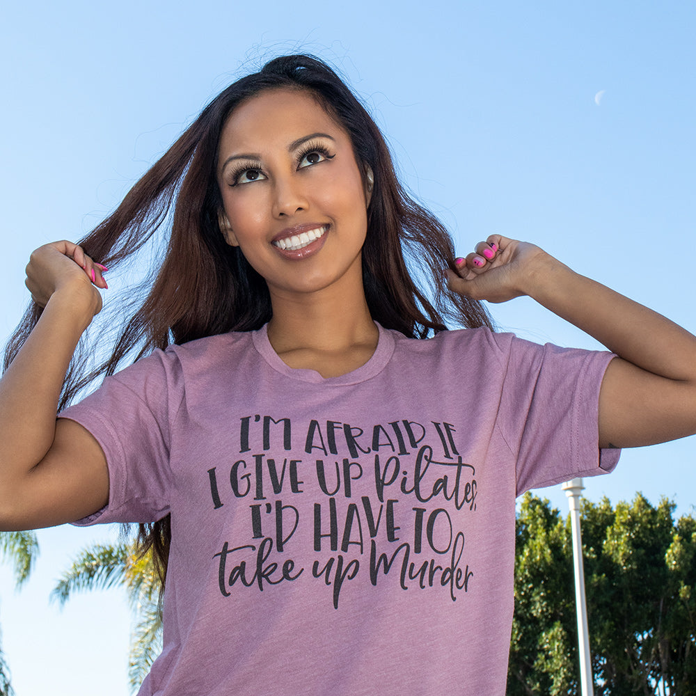 Woman wearing a heather orchid unisex crew neck t-shirt that says "I'm afraid if I give up pilates i'd have to take up murder"