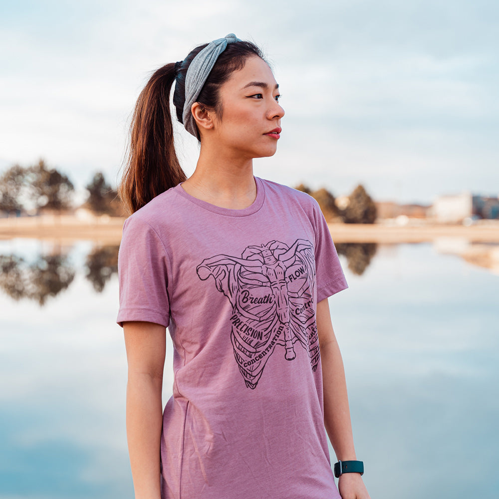 Woman wearing a heather orchid t-shirt with a drawing of a rib cage with the ribs designed as the 6 pilates principles