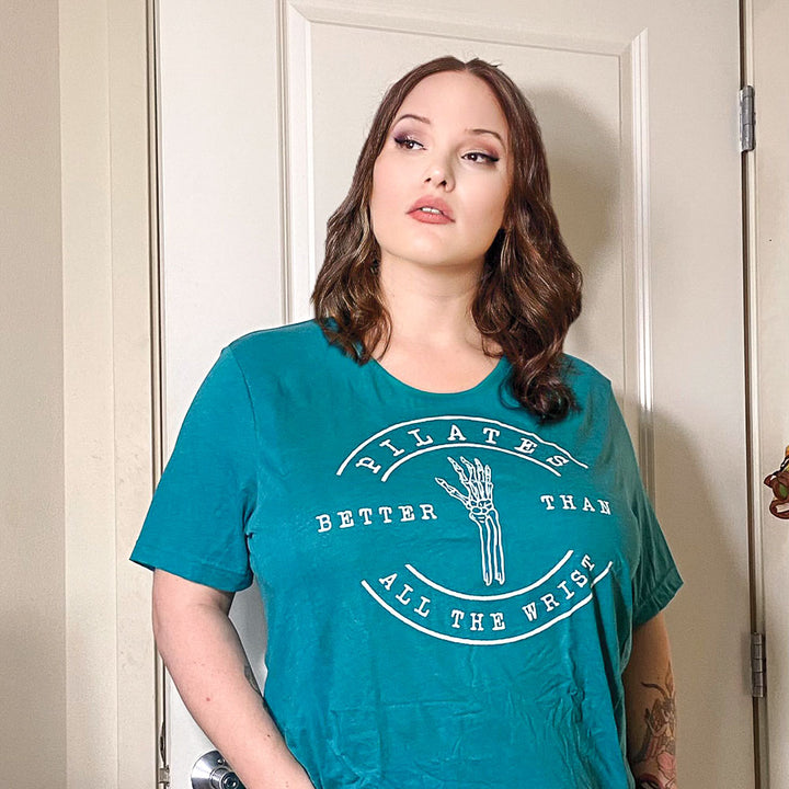 Woman wearing a heather teal unisex crew t-shirt with the words "Pilates better than all the wrist" in white text. There is a skeleton wrist incorporated into the text.