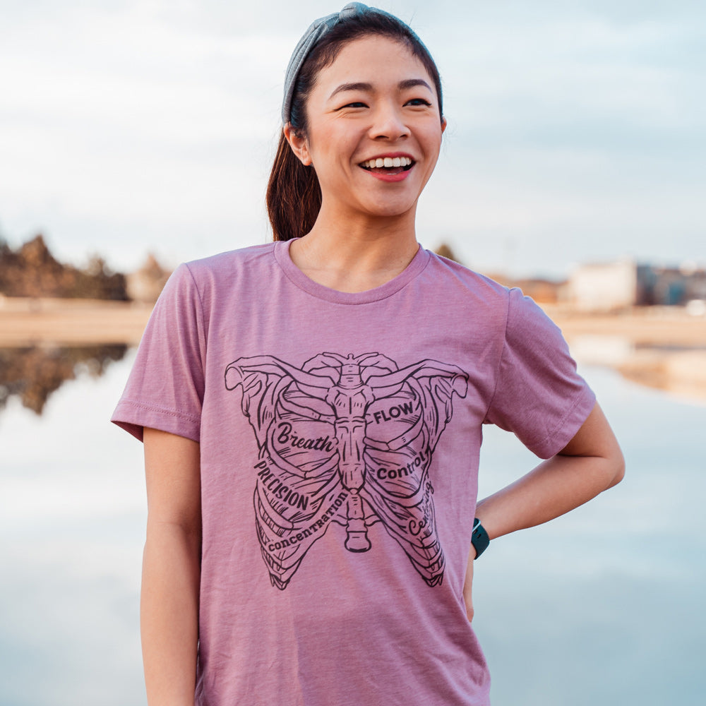 Woman wearing a heather orchid t-shirt with a drawing of a rib cage with the ribs designed as the 6 pilates principles