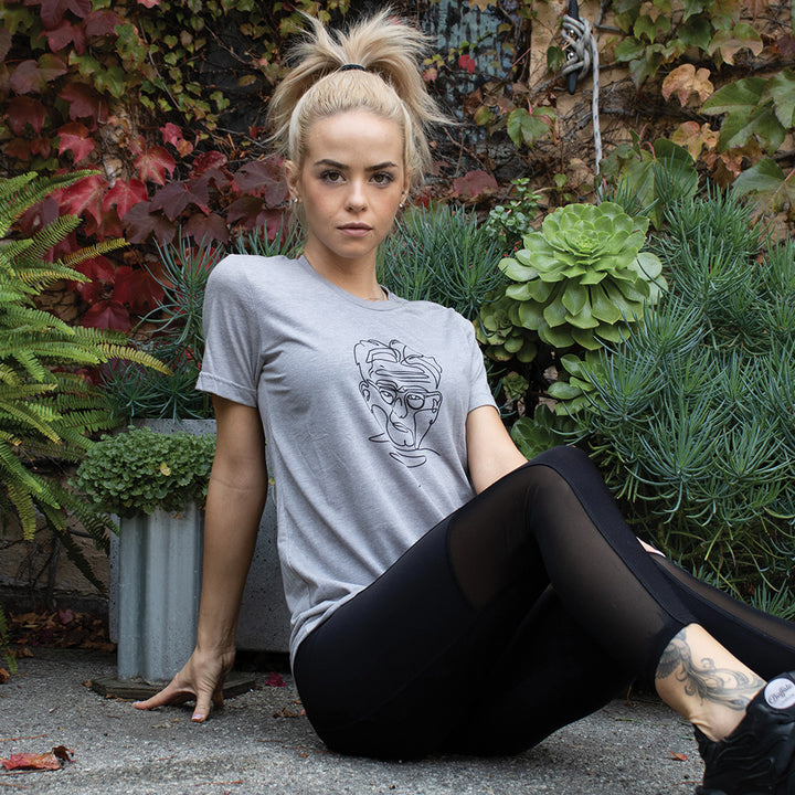 A woman wearing a grey unisex crewneck t-shirt with a line drawing of Joseph Pilates on the front. Joseph Pilates is drawn in black.