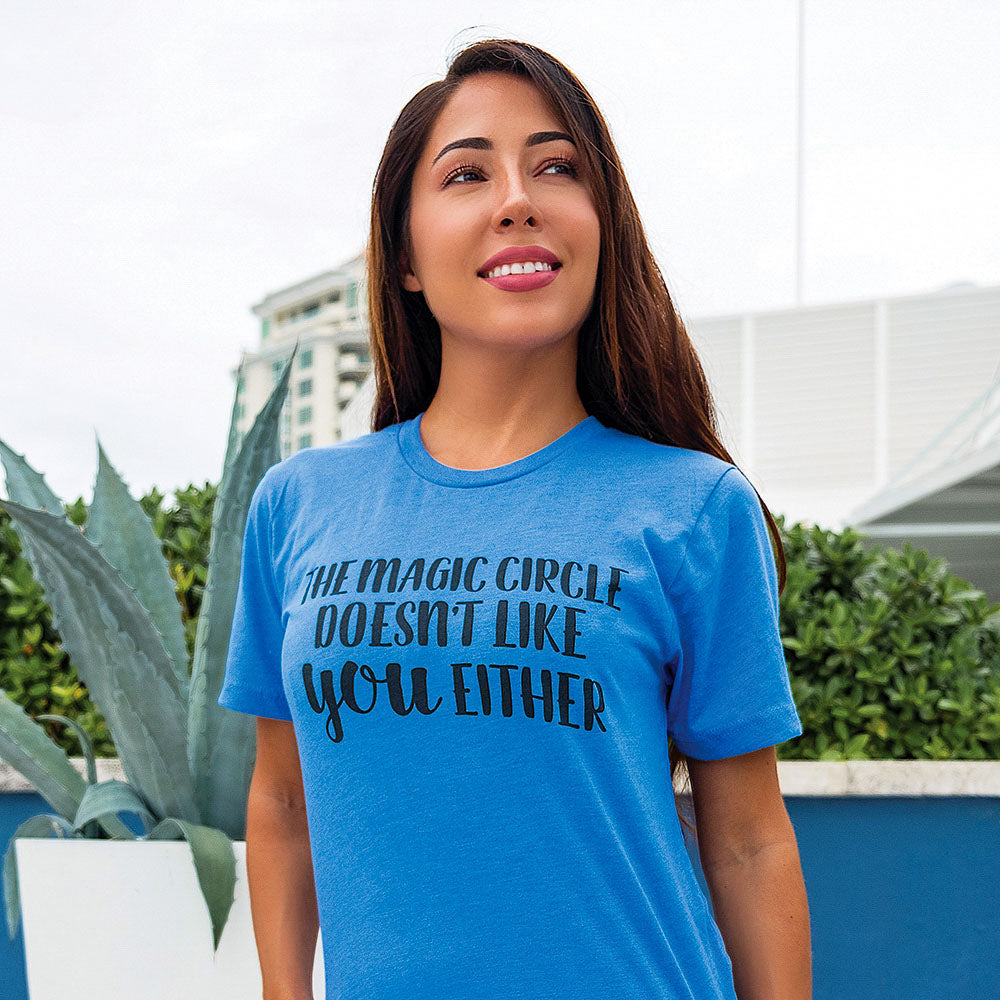 A woman wearing a heather columbia blue shirt that says "The Magic Circle Doesn't Like You Either" in black text