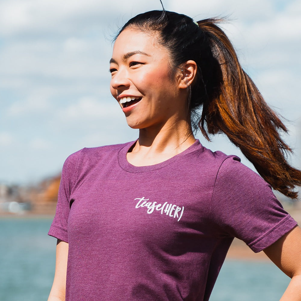 Woman wearing a heather maroon unisex crewneck t-shirt that says "tease(her)" in white text