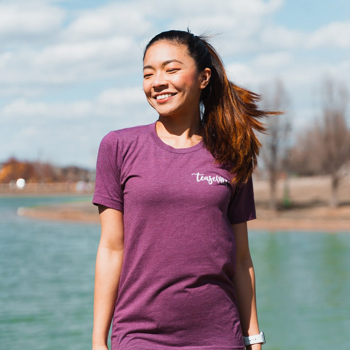 Woman wearing a heather maroon unisex crewneck t-shirt that says "tease(her)" in white text