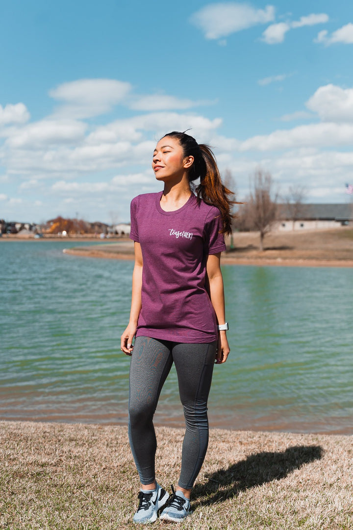Woman wearing a heather maroon unisex crewneck t-shirt that says "tease(her)" in white text