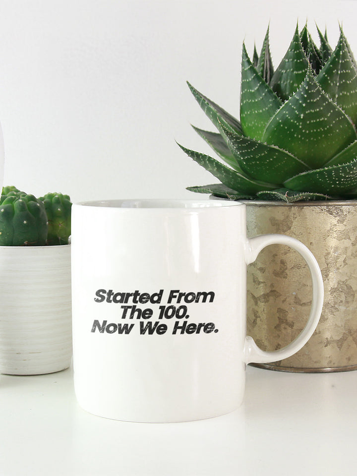 White 11oz coffee mug that says "started from the the 100. Now we here". With plants in the background.