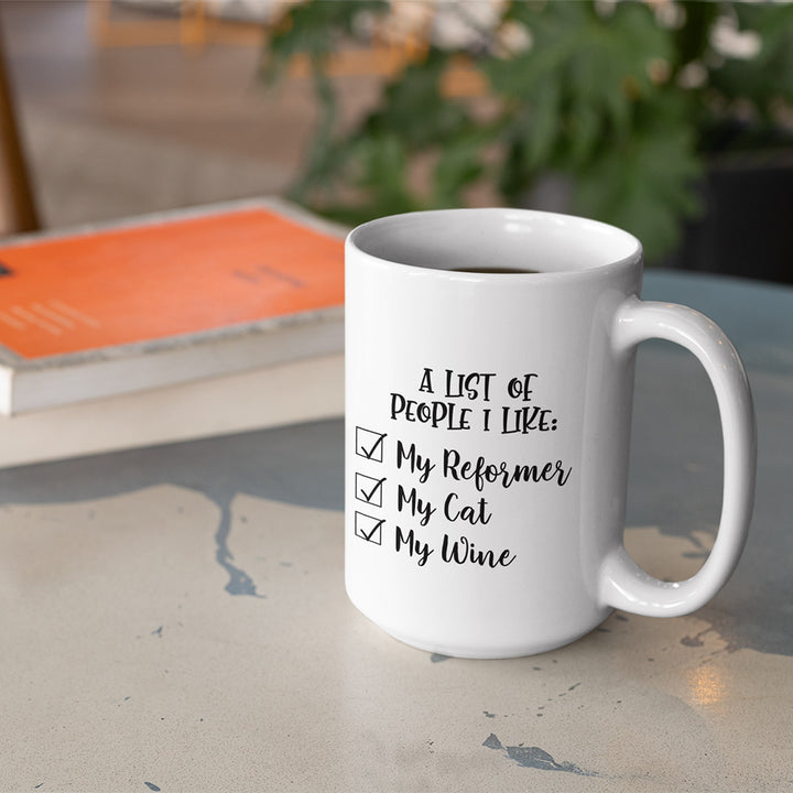 15 oz white coffee mug that says "A list of people I like: My reformer, My Cat, My Wine". Coffee mug is sitting on a marble table with a book in the background.