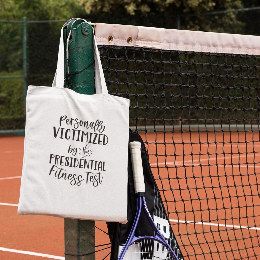 Canvas Tote Bag that says "Personally Victimized By The Presidential Fitness Test". Tote bag is against a tennis bag.