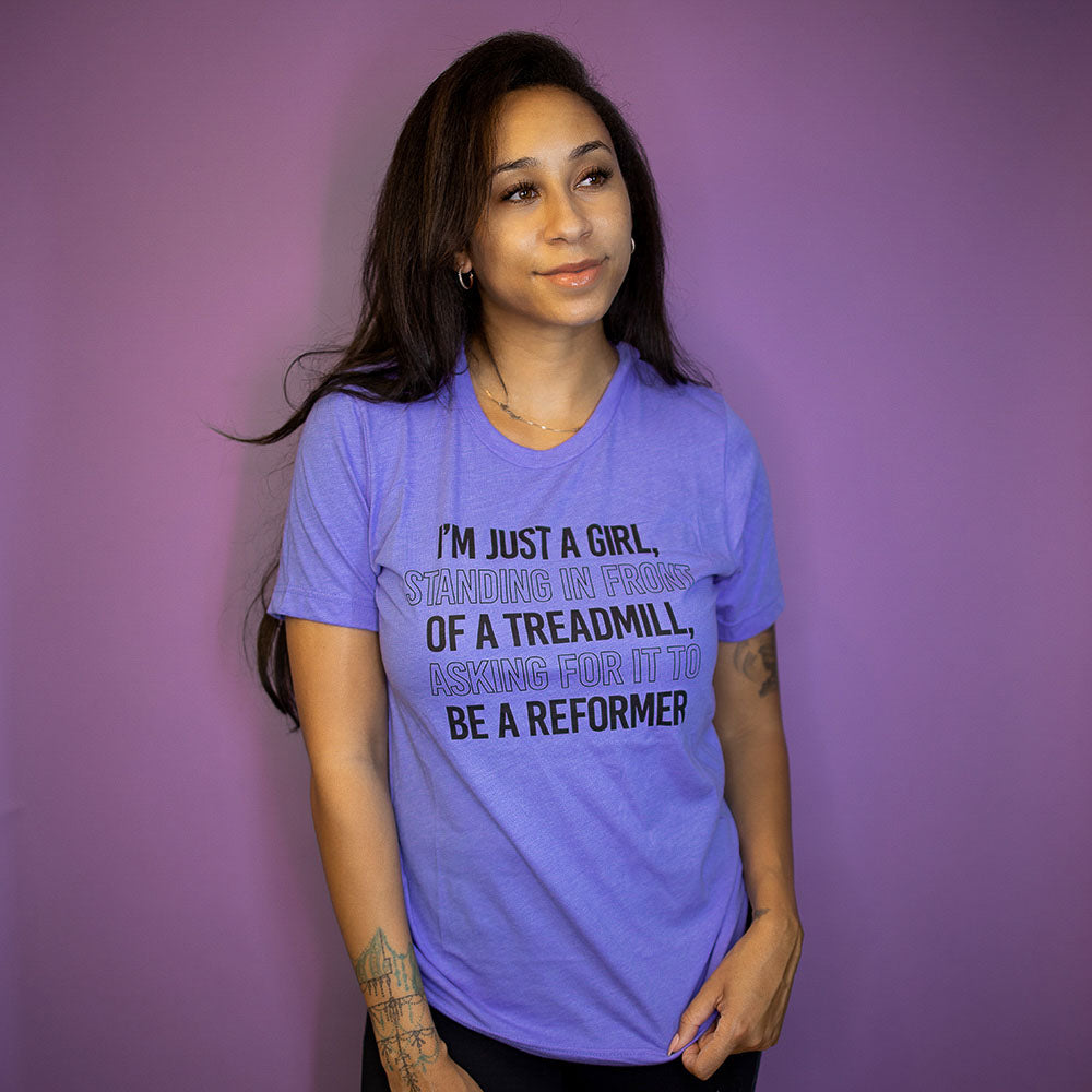 Woman wearing a heather columbia blue crewneck t-shirt that says "I'm just a girl standing in from of a treadmill, asking for it to be a reformer"