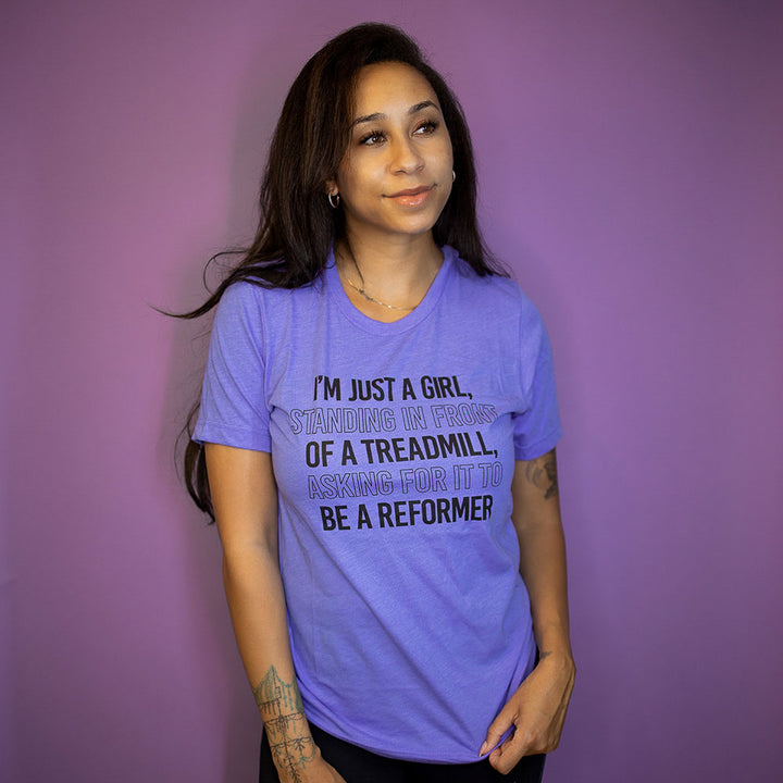 Woman wearing a heather columbia blue crewneck t-shirt that says "I'm just a girl standing in from of a treadmill, asking for it to be a reformer"