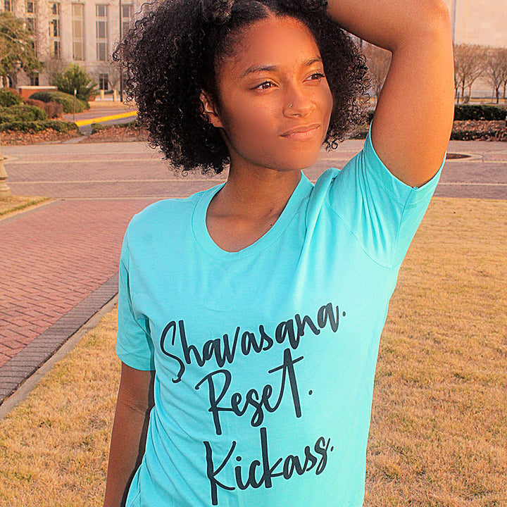 Woman wearing a unisex heather crew t-shirt that says "Shavasana Reset Kickass" in black text