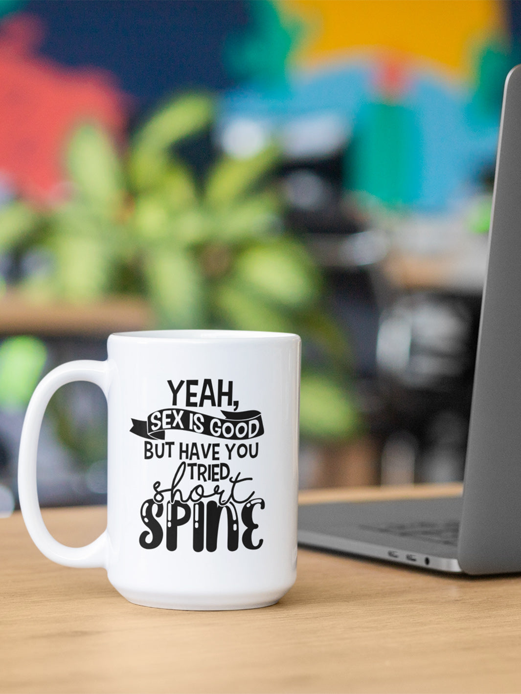 15 oz white coffee mug with handle on the left side. On the mug says " yeah, sex is good but have you tried short spine". In the background is a laptop and blurry plant.