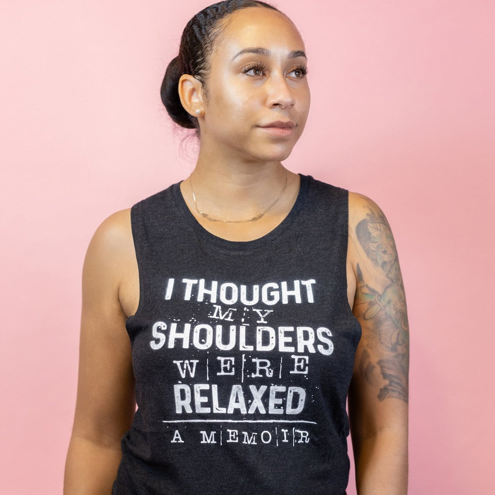 Woman wearing a black muscle tank top that says "I thought my shoulders were relaxed- a memoir" in white text. Woman is leaning against a peach background.