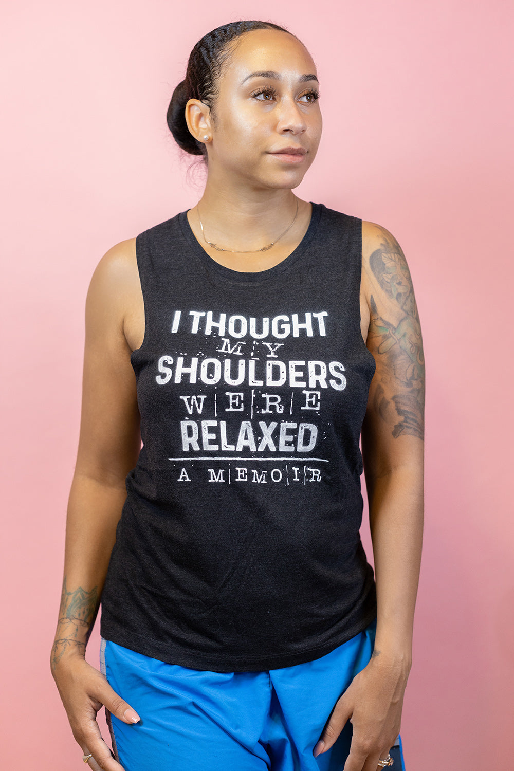Woman wearing a black muscle tank top that says "I thought my shoulders were relaxed- a memoir" in white text. Woman is leaning against a peach background.