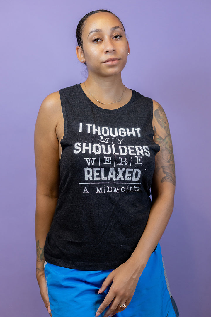 Woman wearing a black muscle tank top that says "I thought my shoulders were relaxed- a memoir" in white text. Woman is leaning a gainst a purple background.