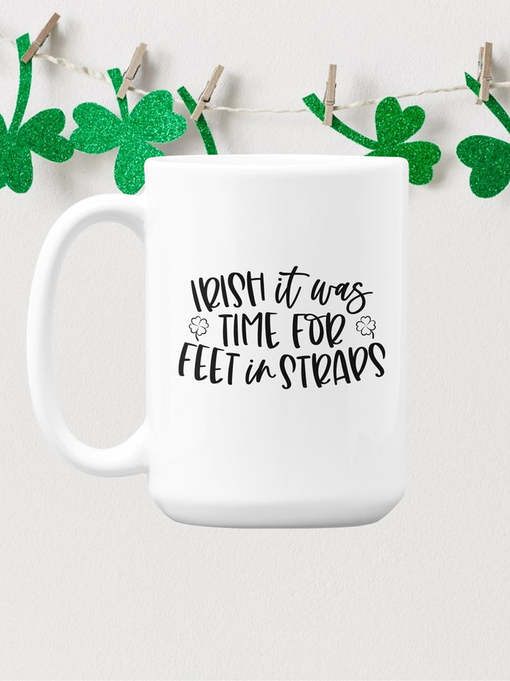 15 oz white coffee mug that says "Irish It Was Time For Feet In Straps" in black text. There are two black outline of 4 leaf clover on the front of the mug. In the background is a banner of green sparkley 3 leaf clover hanging up by clothes pins.